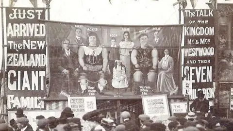 Hull History Centre A black and white photo of people in Victorian dress standing around a circus tent with a poster advertising New Zealand's giant family and a picture of nine people - two of which are extremely large.