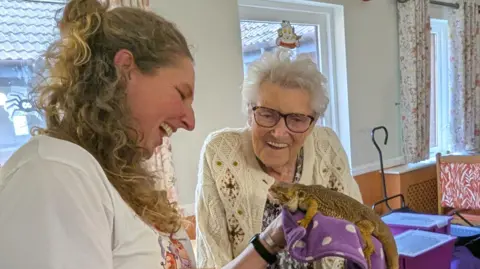 HICA Group Two women are interacting with a bearded dragon lizard. The woman on the left holds the lizard on a purple cloth, while the other lady on the right looks down at it, smiling widely. The lady holding the lizard wears a white t-shirt and has blonde curly hair. The elderly lady on the right has short, white hair, wears large glasses and has on a white knitted cardigan with diamond detailing.