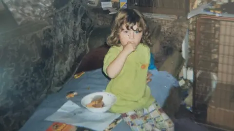 Marcie Reid A young girl with curly brown hair, wearing a green top and patterned trousers, is sitting on the sofa. She has her hand up by her mouth. 