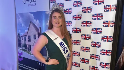 Marcie Reid A woman with long brown hair, wearing a green dress, crown and a sash which says 'Miss York 2025', is posing for a photo. She has one hand on her hip and is smiling at the camera.