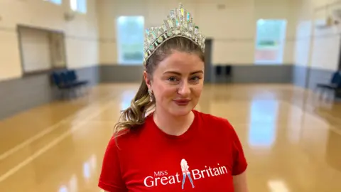 Becki Bowden / BBC A woman brown hair, which is tied back, wears a red t-shirt with the words 'Miss Great Britain'. She is wearing a crown on her head and is smiling at the camera.