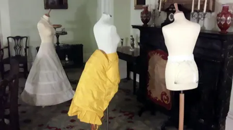 East Riding of Yorkshire Council A historical fashion exhibit with three dresses in a vintage-style room. The first mannequin on the left wears a long, white layered and hooped skirt. The mannequin in the middle has a bright yellow skirt with ruffles on. The third mannequin on the right has a small, white undergarment on. In the background there are chairs and tables, as well as decorative items like candlesticks and vases on top of a fireplace.