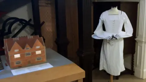 East Riding of Yorkshire Council The image shows a museum exhibit with two displays. On the left, there is a scale model of a brick house with glowing windows, placed on a wooden table with an information plaque in front. On the right, a mannequin is dressed in a long white historical gown with lace sleeves. The background features dark wooden columns and panelling.