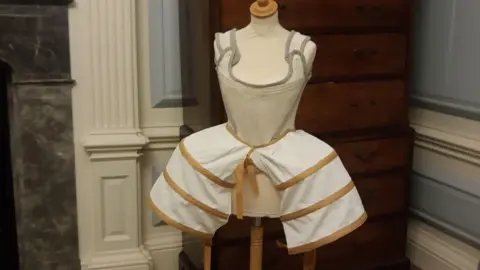 East Riding of Yorkshire Council A museum display featuring a historical undergarment on a mannequin. The garment includes a fitted white bodice with grey trim and shoulder straps, attached to a hooped skirt frame with beige bands around it. Behind the mannequin, there is a wooden chest of drawers and decorative wall mouldings.