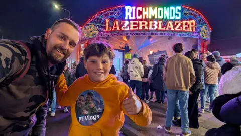 PT Vlogs A man in a camouflaged jumper holding his arm out to take a selfie with his son, a young boy smiling and doing a thumbs up. He is wearing a yellow jumper that says PT Vlogs on it and has another selfie of him and his dad on it. Behind them are people stood around a neon sign which says Richmonds Lazerblazer.