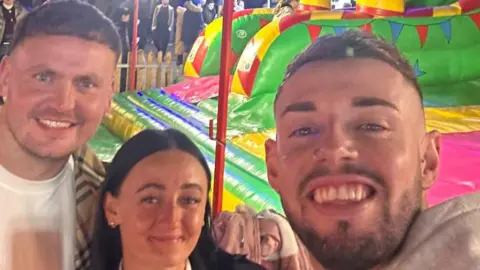 Joel Redhead A selfie taken by a man with short brown hair and shaved sides smiling into the camera and wearing a nose ring next to another men with short brown hair and a woman with long black hair. Behind them is a colourful inflatable.