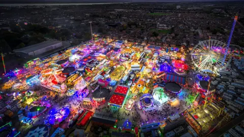 PA Media An aerial view of a funfair with an adornment of neon-coloured lights surrounding a city at night. You can see a Ferris wheel, tall pole-looking rides, swirls and stars.