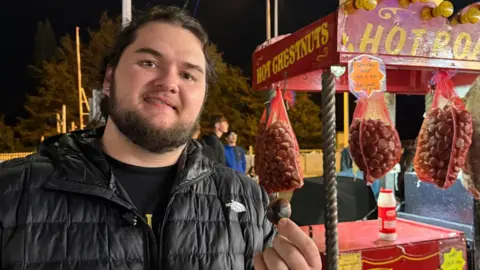 Kalani Ghost Hunter A man with brown hair tied back and a short brown beard smiling into the camera whilst holding a chest nut in his finger. He is wearing a North Face black coat and a vintage red and yellow stall can be seen next to him which says 'hot chestnuts' and has chestnuts in red netted bags hanging from it with a tub of salt.