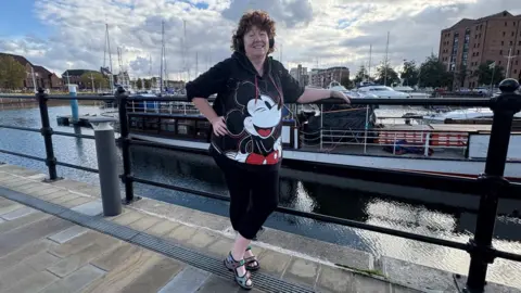 Kevin Shoesmith/BBC A woman in her 60s with brown curly hair is standing on path with a marina and yachts in the background. She is wearing a black T-shirt with a picture of Micky Mouse on it