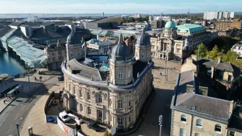A three-sided, grand Victorian building, topped with three domes, at the centre of a city square. To the left, a glass and metal shopping centre stands on stilts above a dock. In the background and to the right, a grand Edwardian building with a large green dome stands on the other side of the square.