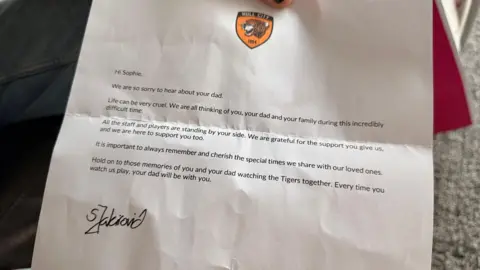 BBC/Jasmine Lowe A letter on white paper. It has a Hull City FC badge at the top. It reads: "To Sophie, We are so very sorry to hear about your dad. Life can be so very cruel. We are all thinking of you, your dad and your family during this incredibly difficult time. All the staff and players are standing by your side. We are grateful for the support you give us, and we are here to support you too. It is important to always remember and cherish the special times we share with our loved ones. Hold on to those memories of you and your dad watching the Tigers together. Every time you watch us play, your dad will be with you."