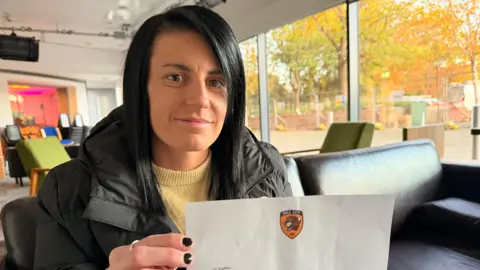 BBC/Jasmine Lowe Sophie Griffiths looking at the camera holding a letter with a Hull City logo at the top. She has long black straight hair with a side parting. She is wearing a black coat and a yellow sweater. Her nails are painted black. She is sitting on a black sofa inside an office-like space. 