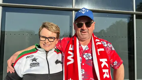Matt Dean / BBC A man dressed in sunglasses, a red Hawaiian shirt, blue baseball cap and Hull KR scarf stands with his arm around his wife, who is wearing a black and white tracksuit and dark glasses
