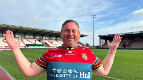 Matt Dean/BBC A man with short brown hair standing by the side of a rugby pitch. He is dressed in a red and green replica kit, with his arms aloft. He is smiling at the camera.