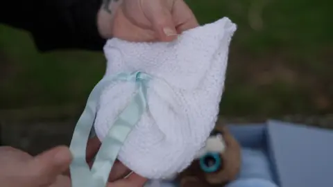 BBC/Joe Weir A small white knitted garment with a green ribbon is held by female hands.