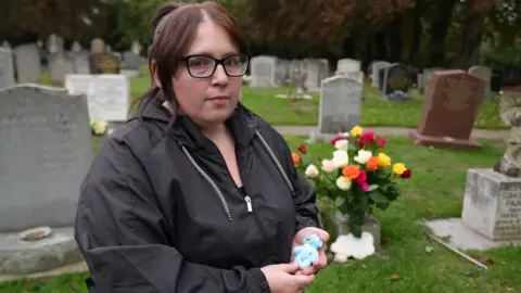 BBC/Joe Bilton Jasmine Beverley wearing a black coat and black glasses holding her premature baby's small blue teddy bear. She is stood in front of her son's grave which is full of coloured roses.