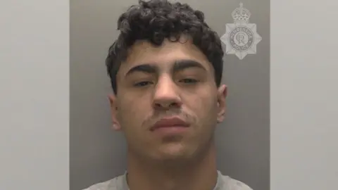 Humberside Police A police custody photo of a man who stares at the camera with a serious expression on his face. He has short, curly black hair and is wearing a light-grey T-shirt. He is pictured in front of a grey background.
