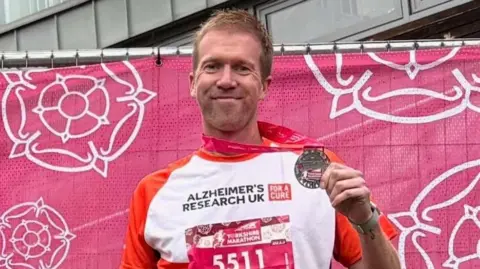 LUKE GLOVER A man holds a silver model proudly, and smiles at the camera. He wears a white and orange t-shirt stating 'Alzheimer's Research UK', and has a pink bib for the Yorkshire Marathon with the number '5511' on. The man stands in front of a pink backdrop with white rose emblems on.