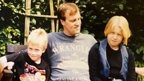 LUKE GLOVER A young boy, older man, and young girl sit on a bench. The older man has his arms outstretched around the children and wears a blue Wrangler t-shirt. The boy has blonde hair and a black graphic t-shirt on, the girl has short blonde hair, a long sleeved black t-shirt and a denim waistcoat over the top. The background of the image features green foliage and a wooden trellis.