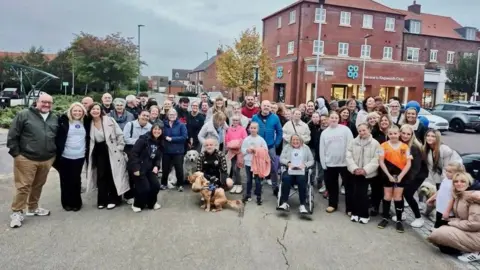 Jenny Duncan A large group of people are stood looking directly at the camera. There is a mixture of males, females, children and pets within the group. Some of them hug, lean on each other and wrap their arms around each other. They are stood in a car park, in front of a red bricked shop with many windows reading: 'Co Op' in blue and 'Welcome to Kingswood's Co op' in smaller white text.