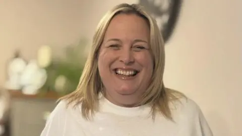 Jenny Duncan A woman is standing indoors, smiling widely at the camera and wearing a white T-shirt. The background is softly blurred, with household items and greenery, such as a clock and plants. She has blonde hair just past her shoulders.