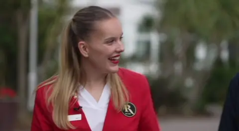 A woman with blonde hair, tied back in bunches, laughs at something out of frame on the right. She is wearing a red jacket, white shirt and red lipstick.