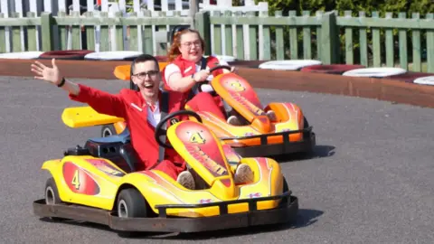 Stephen Gamage A young man beams widely at the camera has he drives a yellow go-kart around a track. He is waving with one arm towards the camera. He is wearing a red jacket and trousers. To the right, a woman with ginger hair and glasses, also dressed in red, drives a similar car. 