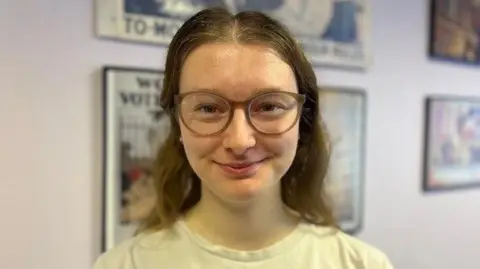Amelia Bateman-Young is smiling into the camera. She's wearing glasses and a white t-shirt and has long brown hair. She's standing in front of a wall with pictures hanging from it.