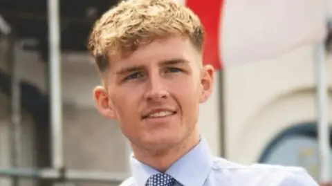 Oliver Freeston Oliver Freeston is smiling into the camera. He has blonde curly hair and is wearing a white shirt with a blue and white tie. He's stood in front of a building with a flag flying behind him.