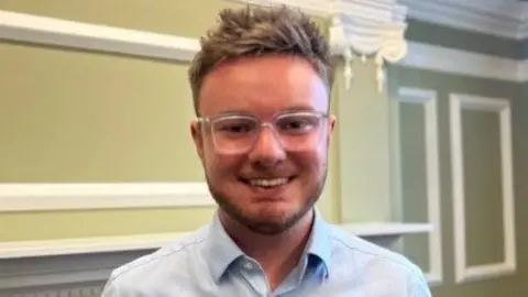 Ted Dolman smiling into the camera. He has brown, short, spiky hair and is wearing glasses. He's also wearing an open collar blue shirt. He's standing in an office within Hull's Guildhall.