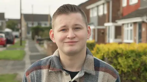 BBC Frankie Williams with short brown hair smiling into the camera. He's wearing a blue and cream checked shirt. He's stood outside in a residential area and in the background are houses, a hedgerow and parked cars.