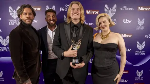 Handout Pete Wicks, who has brown hair and facial hair wearing a full brown velvet suit. Sam Thompson is standing next to him wearing a navy suit and a white shirt. Marcus is standing next to him wearing a full black tuxedo holding a trophy. He has shoulder-length blond hair. On the far right is Anne Marie with a short blonde bob, wearing a large gold necklace, a black corset and a black skirt. There are all standing in front of a purple wall with Pride of Britain, ITV and Daily Mirror branding.