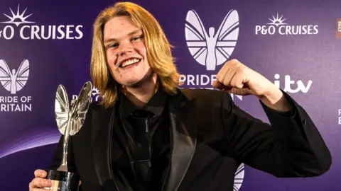 Handout Marcus with shoulder-length blond hair wearing a full black tuxedo smiling at the camera. He is holding a silver trophy and is holding his left arm up in a celebratory fist motion. He is stood in front of a purple wall with branding on.