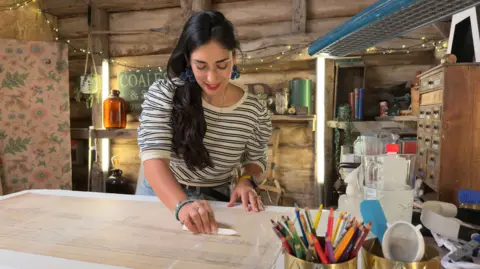 Ricochet Ltd Angelina Bakalarou stood over a white table working on repairing a large drawing of the Humber Bridge. She has long black hair and is wearing a striped long-sleeved top and blue jeans. She is in a wooden shed surrounding by materials for restoring paper. 