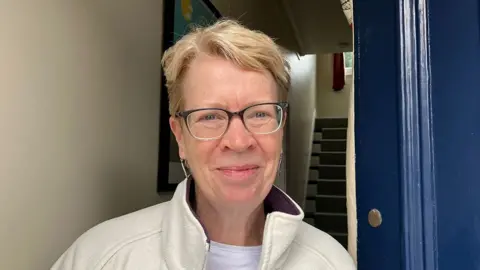 Amanda White / BBC Kate Sunley standing by the door of a house she has short blond hair and wears glasses and a white shirt and cream top
