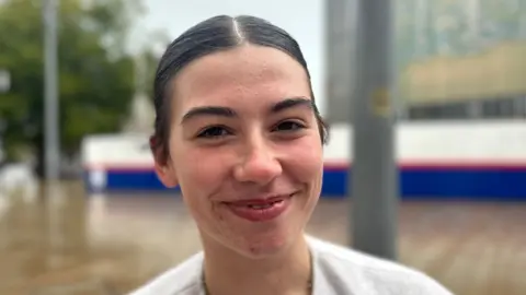 BBC News A girl with dark her and eyebrows, she's smiling in the middle of Hull city centre and her hair is slick back. It's in a pony tail and it matches her dark brown eyes. She looks in her early twenties. 