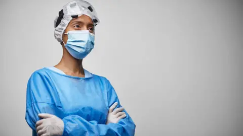 Getty Images Angled view of young female doctor in hospital workwear, bouffant cap, surgical mask and protective eyewear looking away with a serious expression. 