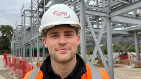 Louis Sangwin, wearing a white hard hard and orange high-vis coat and is standing in front of a steel frame on a building site.