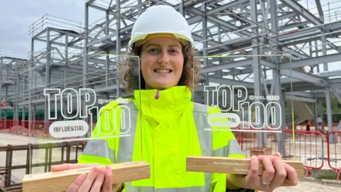 BBC Katy is wearing a broad smile, a white hard hat, and a fluorescent yellow jacket as she holds up two awards, one in each hand. She is standing on a building site with a steel construction frame behind her.