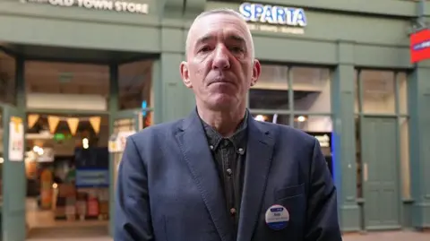 Crispin Rolfe / BBC Rob Ellis standing in front of his shop. He has short hair and is wearing a dark blue jacket and shirt 