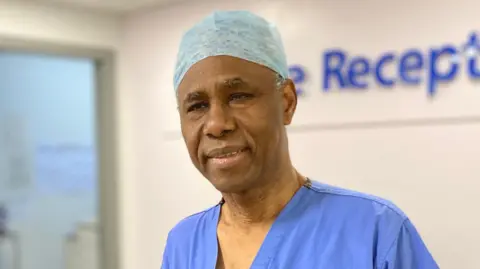 BBC Mr Akomolafe is stood in front of a hospital reception desk. He is wearing theatre scrubs, which includes a light blue surgical cap and blue top.