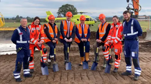 A group of 8 men and women standing in a muddy field in front of a helicopter. They are wearing high vis jackets and a number of them are holding spades.