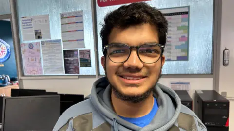 A man with glasses, smiling at the camera. He has a blue top on with a grey pull string jumper. His glasses are circular and large on his face, he's stood in front of a series of stacked up recycled PCs and looks excited to be talking on the matter 