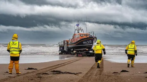 Mike Milner RNLI Three RNLI crew members in orange, black and yellow all-weather gear walk on a sandy beach towards an orange and black lifeboat as it is towed into the waves beneath a dark, stormy sky.