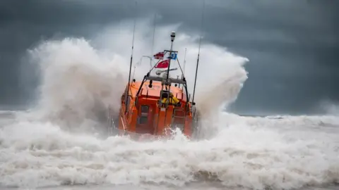 Mike Milner RNLI An orange RNLI lifeboat seen from the back launches head-on into heavy seas, causing a huge bow wave to spray up at either side of the vessel. White foaming waves crash in the foreground and a dark, stormy sky forms the background.