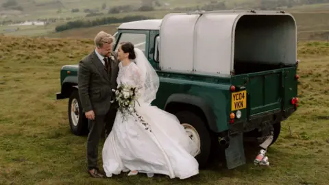Joe Hall Photography A newly-married couple pose in front of a dark green Land Rover Defender that is parked on a grassy hill. The bride wears a floor-length white gown with lace details and a short veil. She holds a bouquet of white and purple flowers. The groom wears a dark-coloured tweed suit and purple tie.