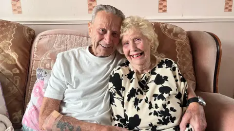 BBC Harry and Rose Brumhead smile at the camera while hugging each other on the sofa with their heads against one another. Harry has grey hair and is wearing a white t-shirt. Rose has blonde, curled hair and is wearing a cream-coloured jumper with black flowers.