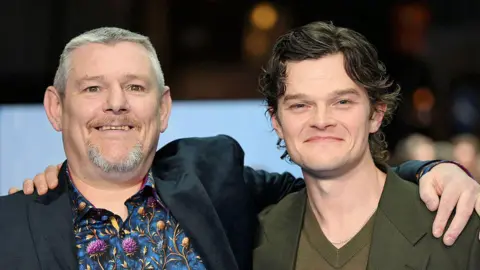 Getty Images John Davidson (left) and Robert Aramayo (right) stood arm-in-arm looking at the camera. John Davidson has short grey hair and short grey facial hair. He is wearing a blue patterned shirt and a black blazer. Robert Aramayo has short curly brown hair is wearing a khaki v-neck sweater and a khaki blazer. The background behind them is blurred. 