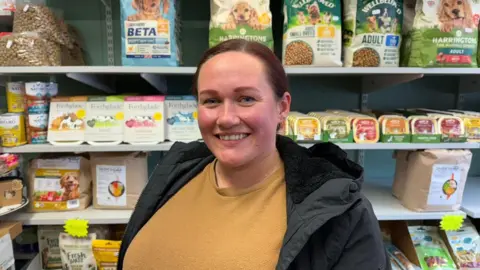 BBC/ Grace McGrory The photo shows a woman smiling at the camera. She's wearing a mustard coloured jumper, and a black coat. She has brown hair which is tied back. She's standing in front of shelves, which contain a variety of animal food. 