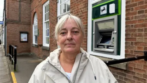 BBC/ Grace McGrory The photo shows a woman looking at the camera. She is wearing a cream jacket, and has white hair. She's standing in front of a red brick building. To her right, there is a cash machine with the Lloyds logo on it. 